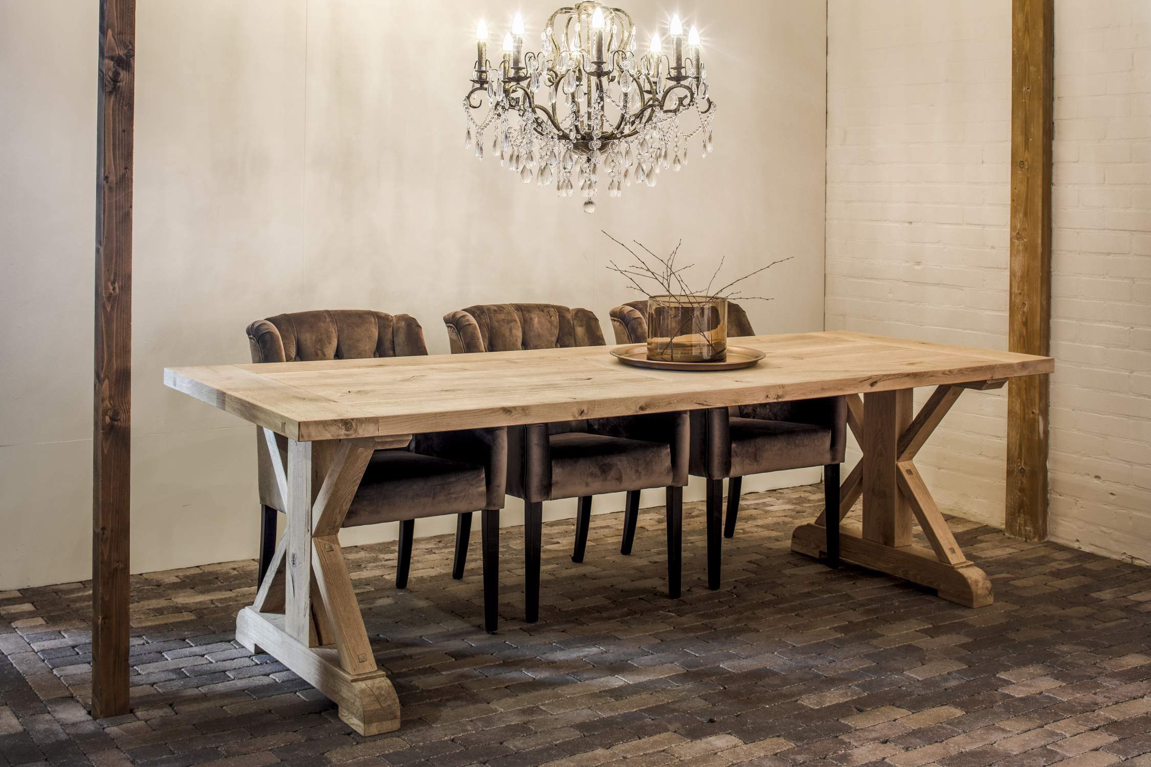Long dining table made of light wood with an X-frame, brown upholstered chairs and a crystal chandelier.