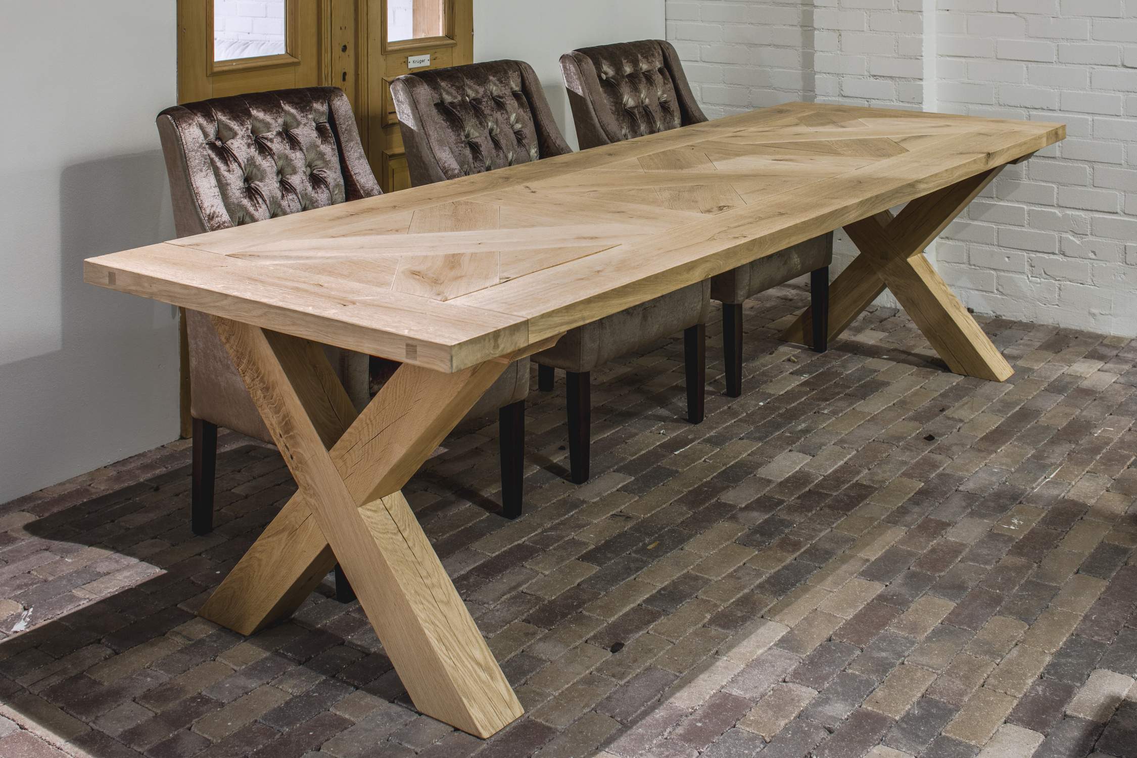 Long dining table made of light wood with an X-frame and upholstered chairs on a brick floor.