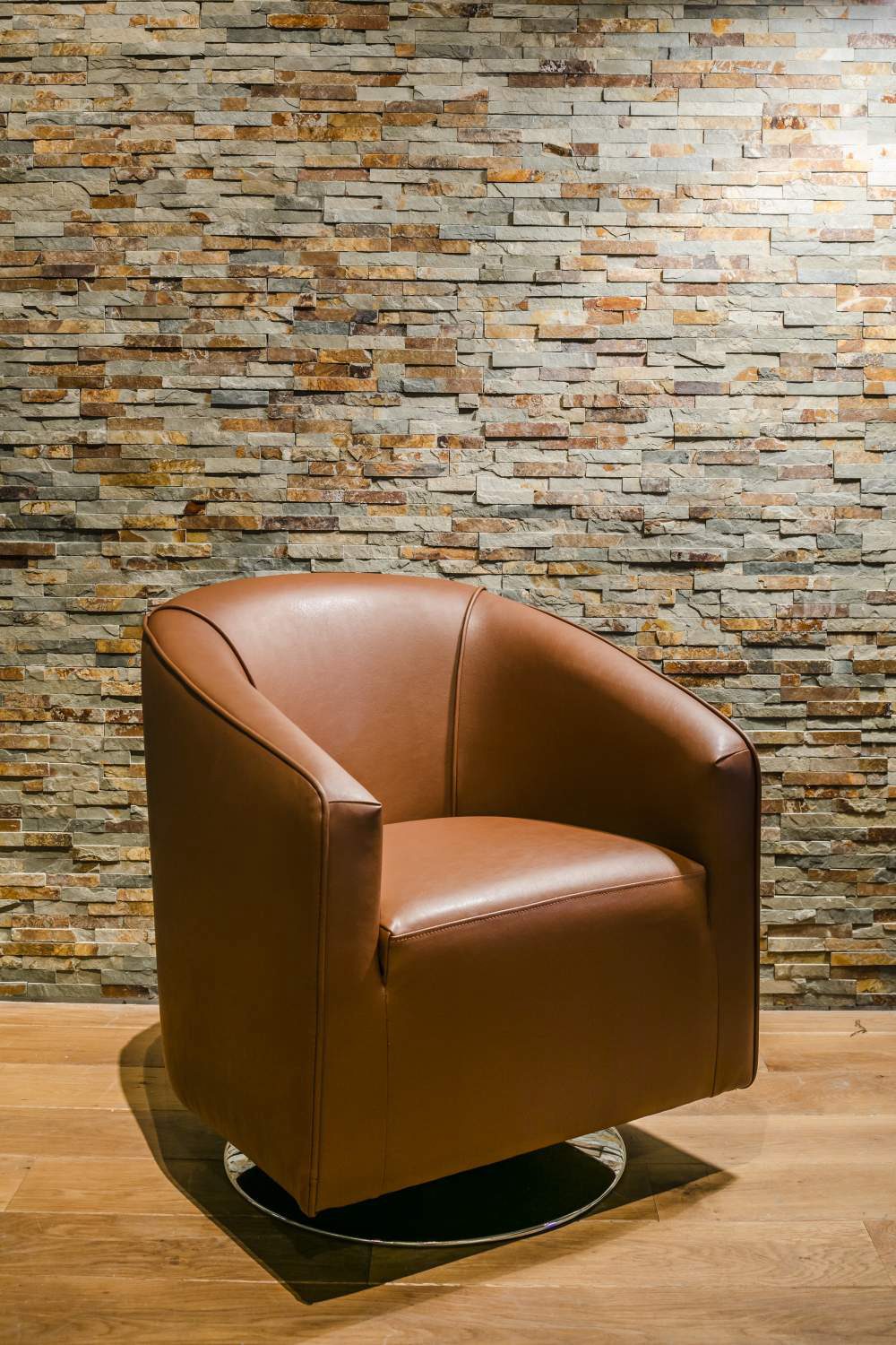 Poltrona girevole color cognac con base rotonda davanti a una parete in pietra naturale su pavimento in legno.