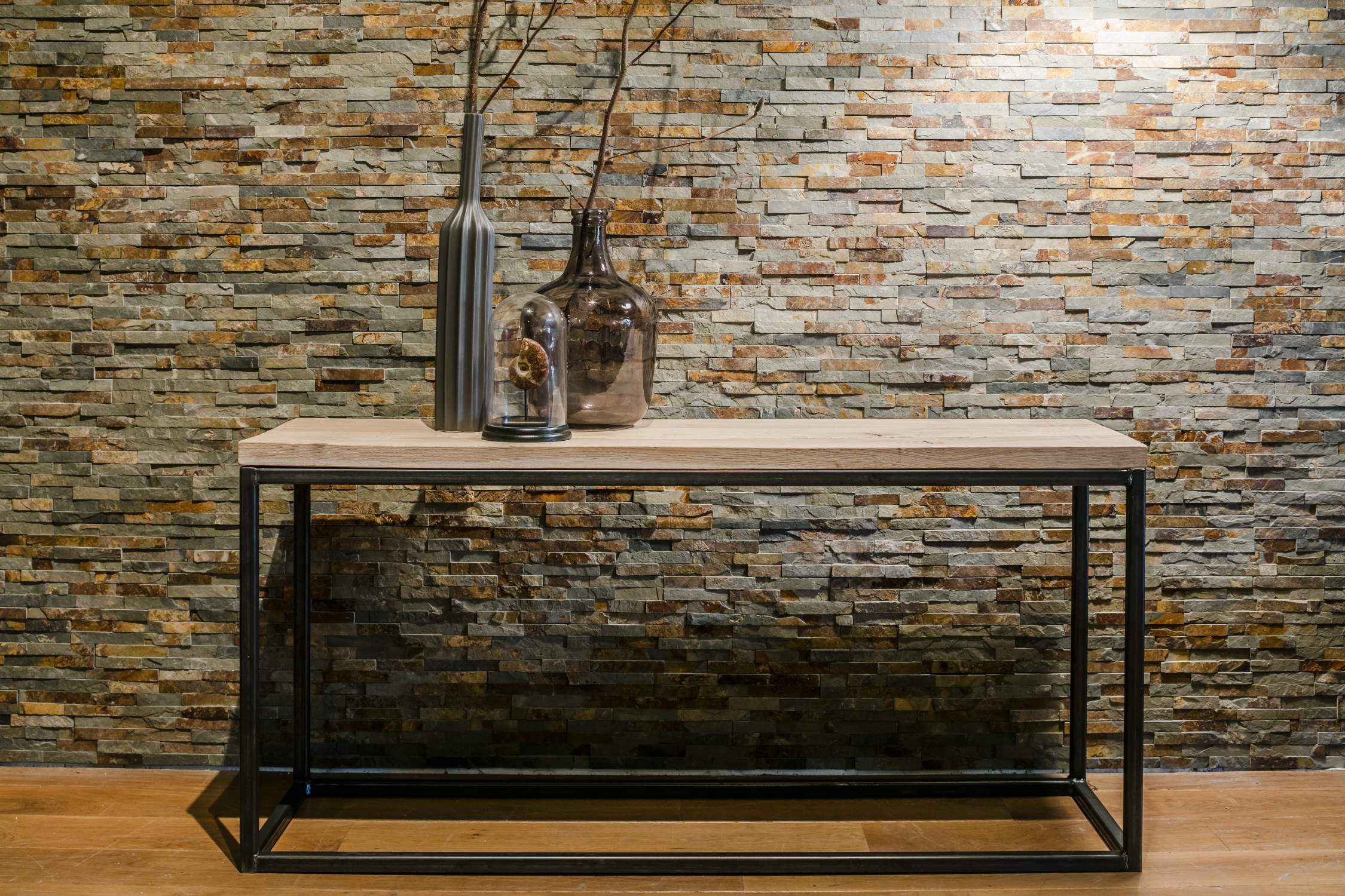 Long console table with a light wooden top and black metal frame in front of a natural stone wall.