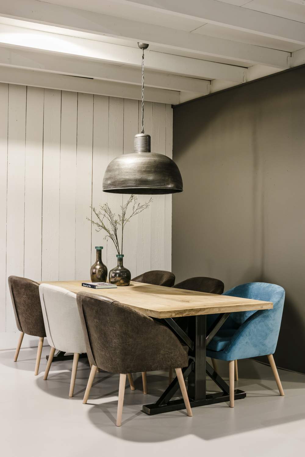 Light wooden table with a black metal frame, surrounded by coloured upholstered chairs under a large pendant light.