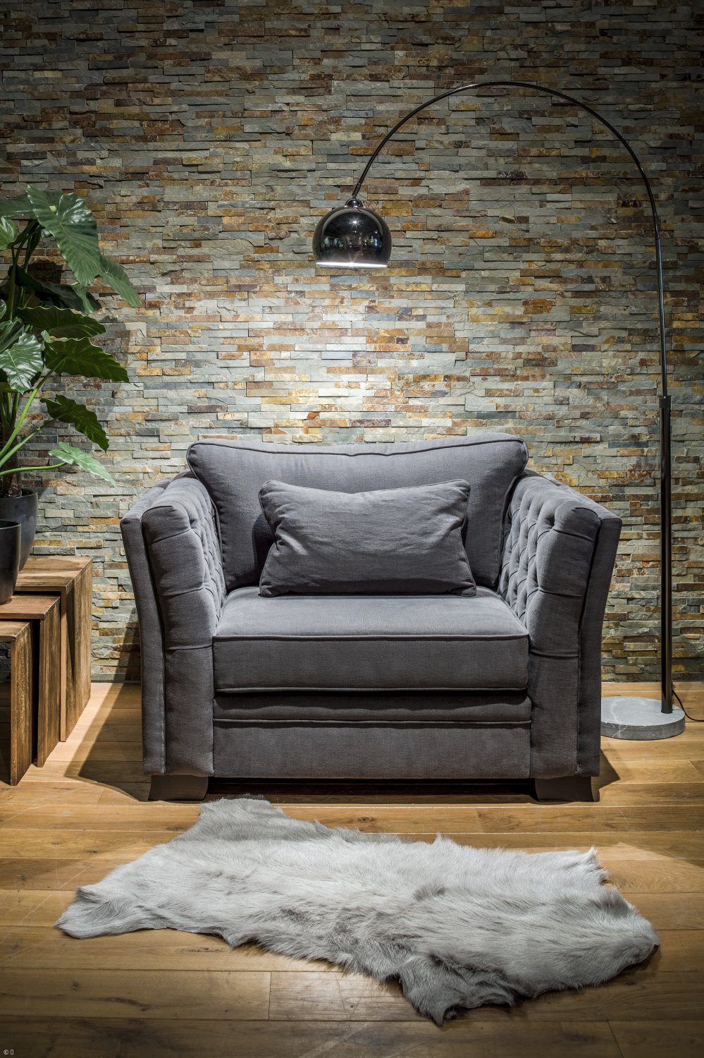 Dark grey upholstered armchair with a cushion in front of a natural stone wall and a grey fur rug.