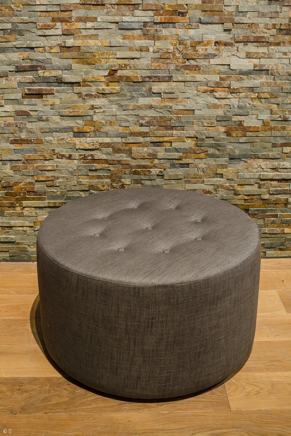 Round dark grey upholstered footstool with button tufting on a wooden floor in front of a natural stone wall.