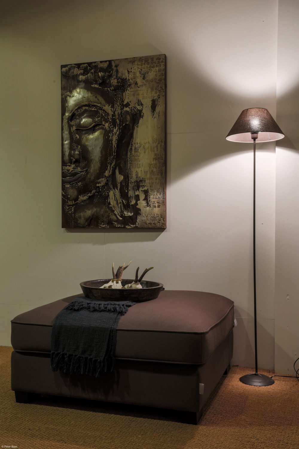 Brown rectangular upholstered footstool with a dark throw and bowl next to a slender floor lamp.