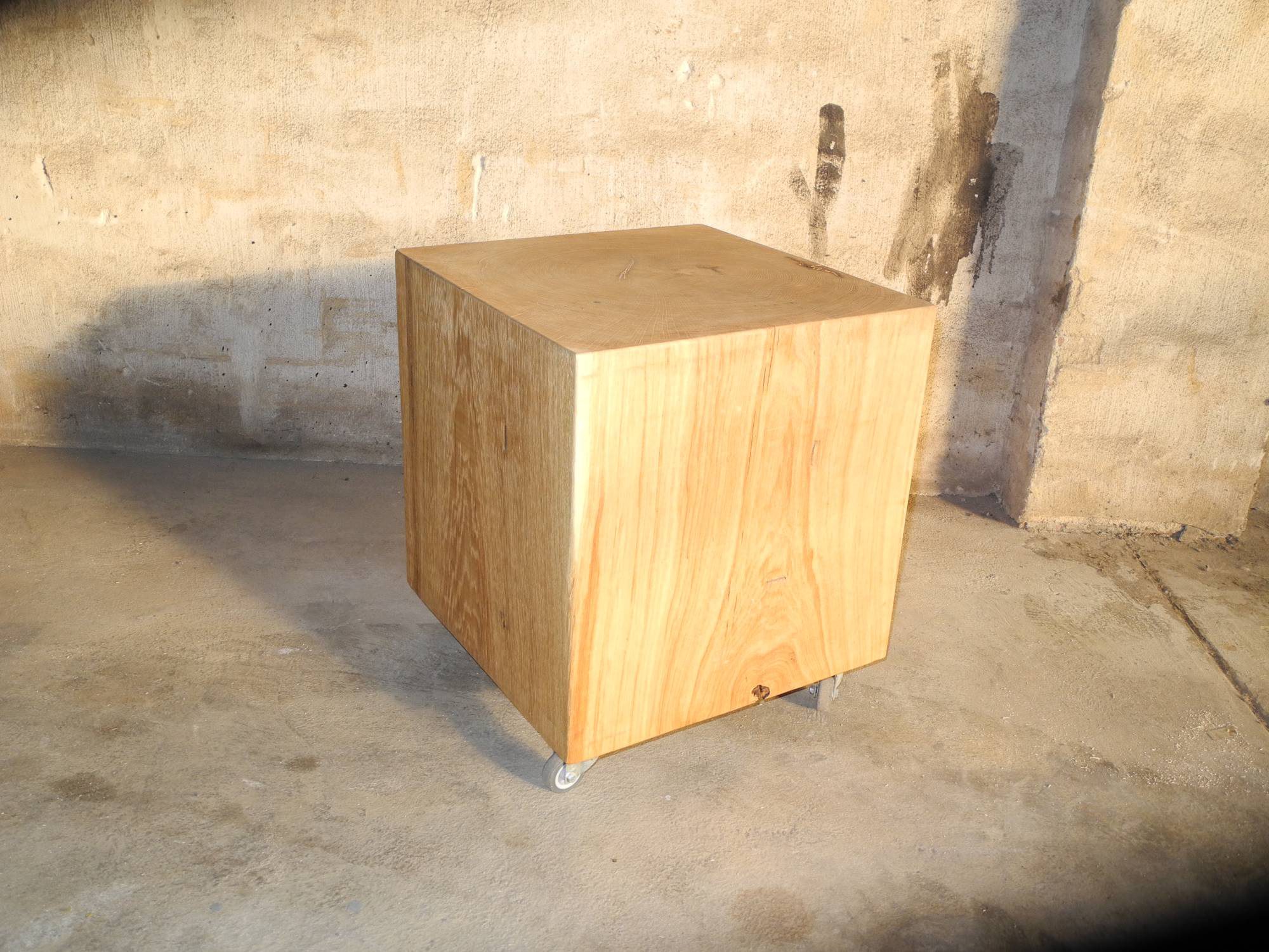 Warmly lit solid wood cube on castors in front of a mottled concrete wall, casting a shadow.
